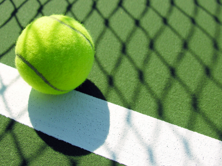 A tennis ball lying on the line. The shadow of the net is being cast across the ground.